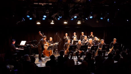 L'Orchestre de jazz d'Ottawa se produit sur une sc&egrave;ne faiblement &eacute;clair&eacute;e, o&ugrave; les musiciens jouent divers instruments, notamment le piano, la contrebasse, la batterie, les saxophones, les trompettes et les trombones.