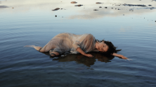 Personne allong&eacute;e dans de l&rsquo;eau peu profonde pr&egrave;s du rivage, partiellement immerg&eacute;e et portant un v&ecirc;tement l&eacute;ger et translucide, avec des rochers dispers&eacute;s dans l&rsquo;eau calme. © Jade Gabriele