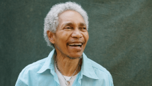 Personne aux cheveux gris courts portant une chemise bleu p&acirc;le et un collier, tourn&eacute;e l&eacute;g&egrave;rement de c&ocirc;t&eacute; avec la bouche ouverte, devant un arri&egrave;re-plan vert textur&eacute;. © Wade Muir