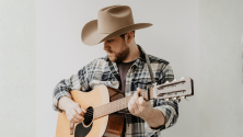 Personne portant un chapeau &agrave; large bord et une chemise &agrave; carreaux jouant de la guitare acoustique sur fond clair. 