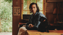Personne assise dans une cabane en bois pr&egrave;s d&rsquo;une fen&ecirc;tre, avec de la verdure &agrave; l&rsquo;ext&eacute;rieur, tendant la main vers un petit chat noir sur une table en bois; des livres, une lampe et des affiches sont visibles dans l&rsquo;int&eacute;rieur chaleureux. © Jodi Heartz